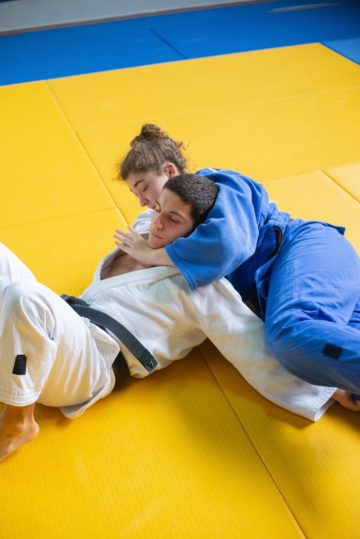 Two adults practicing judo in a grappling hold on a yellow mat indoors.